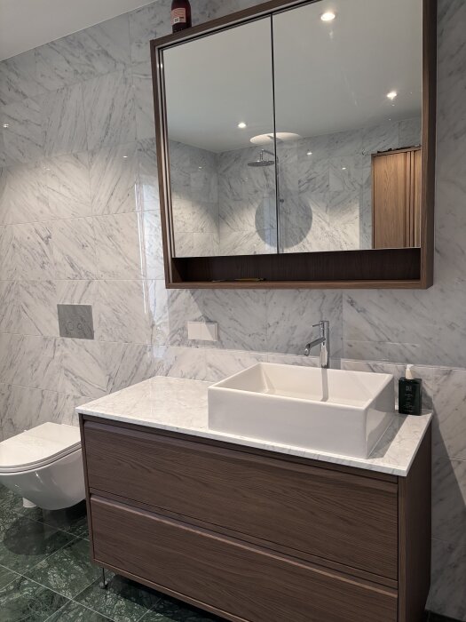 Badrumsinstallation med vit bänkskiva, monterad blandare och handfat, träskåp och spegel med marmordetaljer på väggarna.