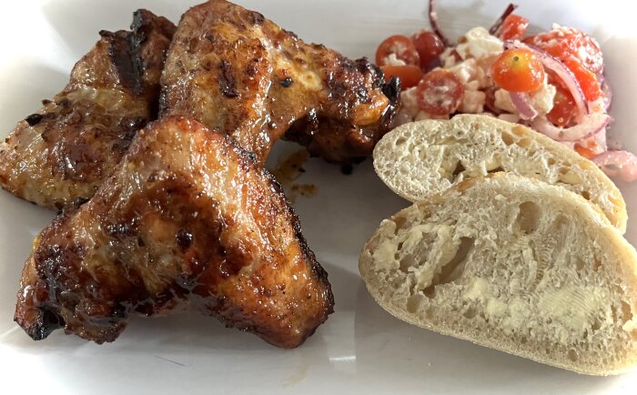 Grillade kycklingvingar med surdegsbröd och fetasallad med tomat och lök på en vit tallrik.