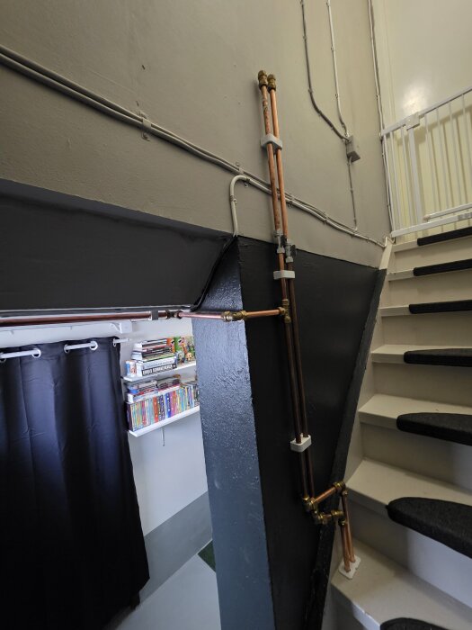 Källartrappa med nylagda kopparrör och svarta trappstegsmattor, hylla med böcker och en gardin i bakgrunden.