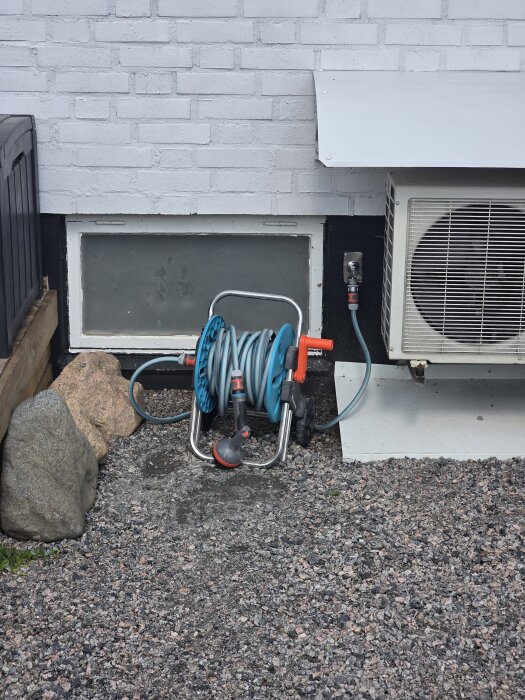 En trädgårdsslang på rulle med ansluten vattenkran utomhus på en stenlagd yta framför ett vitt tegelhus med luftkonditionering.