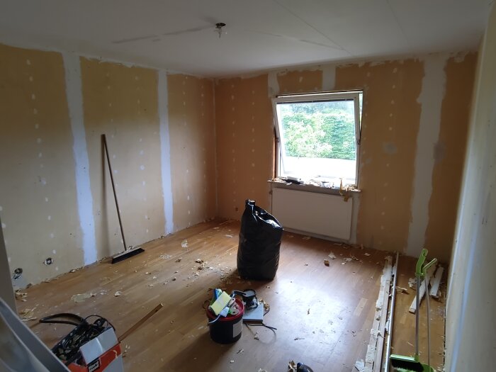 Rum under renovering med avskalade väggar, verktyg, och en sopsäck på golvet. Fönster med utsikt över grönska.