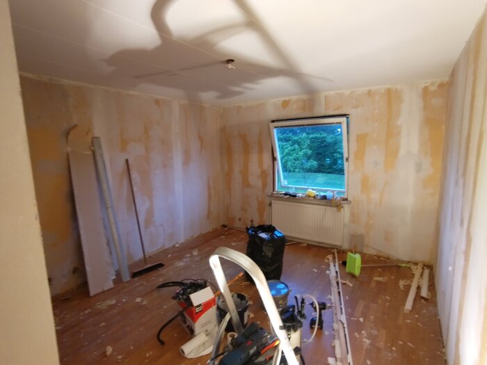 Rum under renovering med borttagen tapet, verktyg på golvet och utsikt mot en grön gräsmatta genom fönstret.