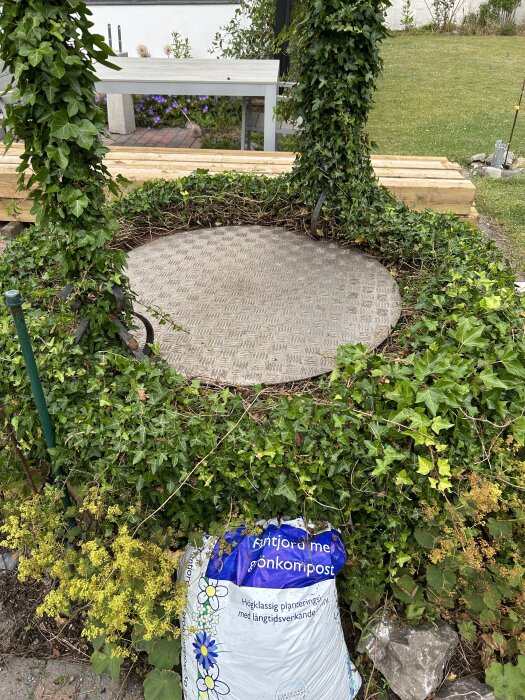 Rund brunn med durkplåt omgiven av murgröna. På marken ligger en säck med jord och stenar, en trädgårdsmöbel syns i bakgrunden.