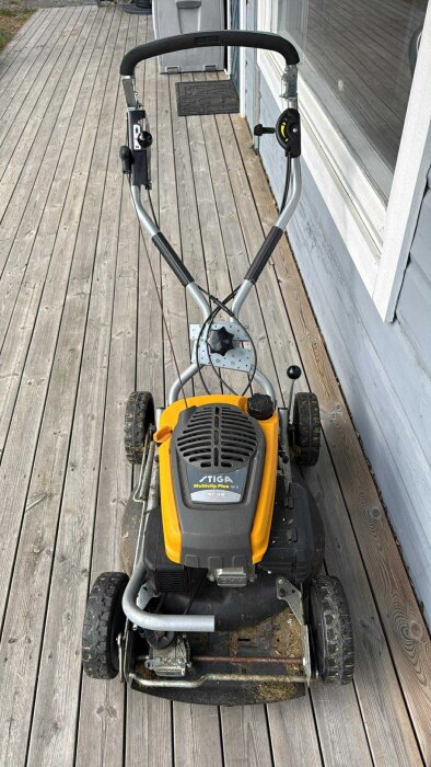 En gul och svart Stiga Multiclip Plus 50 S gräsklippare placerad på en altan i trä.