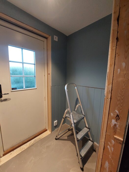 Blåmålade väggar, trädörr med fönster, stege och omålade foder i ett rum. Golvet saknar klinkers och rummet är delvis under renovering.