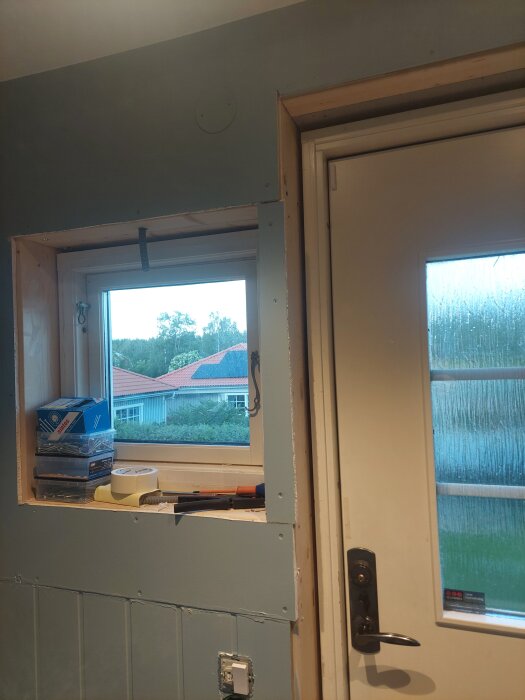 Färdigmålad vägg med ett fönster och dörr utan lister. Verktyg och tejp ligger i fönsterkarmen, redo för fortsatt renovering.