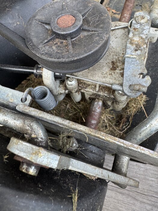 Närbild av gräsklipparens remskiva och fjädersystem, delvis täckt av gräs och smuts.