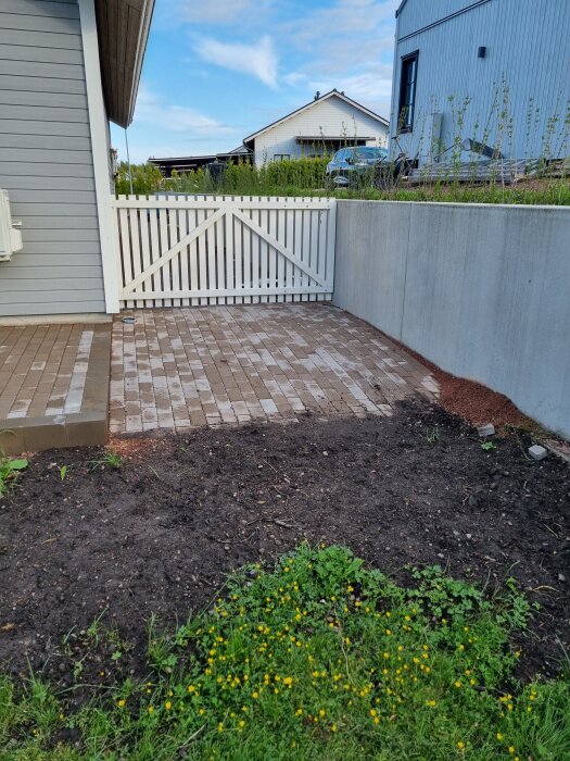 Stenbelagd yta vid sidan av ett hus med en vit grind och en mur av betong. Ogräs och gulblommig växtlighet syns i förgrunden.