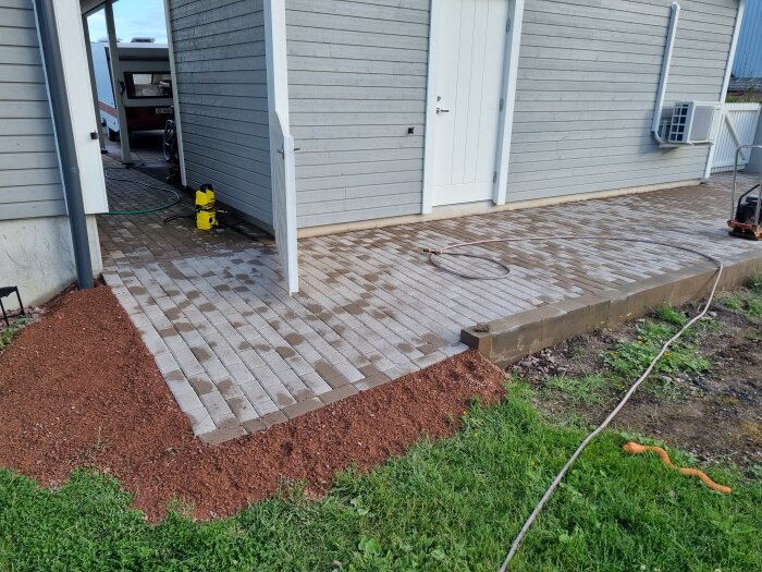 Ny stenläggning framför ett hus med sluttande kant, omgiven av jord och gräs; trädgårdsslang syns på marken.