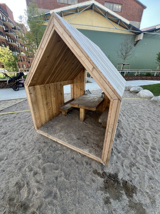 Litet trähus på lekplats, byggt i enkelt snedtak, med sittplats och bord inuti. Bakom syns en stor byggnad och grusunderlag runtom.