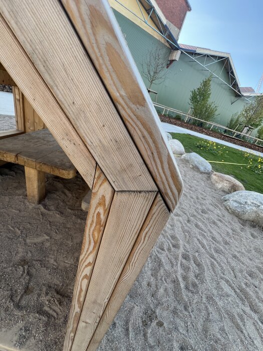 Vinkel av träkonstruktion, närbild av snedställda träplankor i parkområde med stenar och sand, grönt plåttak i bakgrunden.