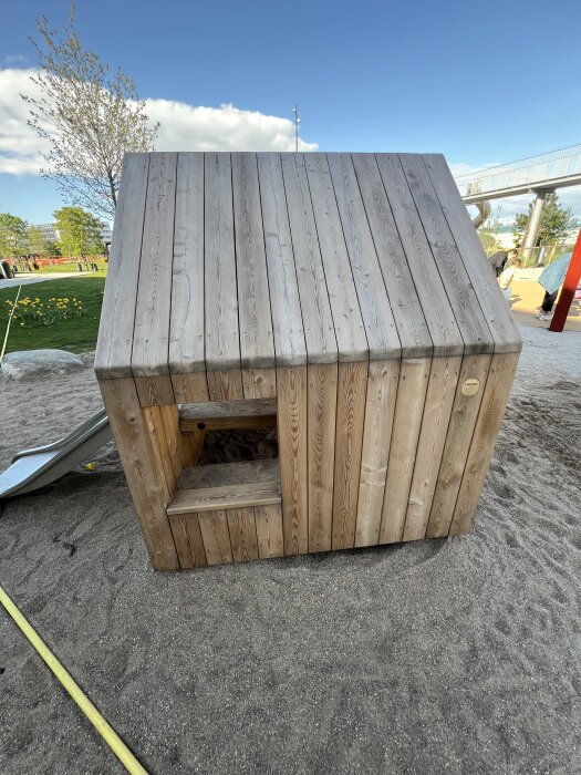 Trälekstuga med sluttande tak, omgiven av sand och grönska.