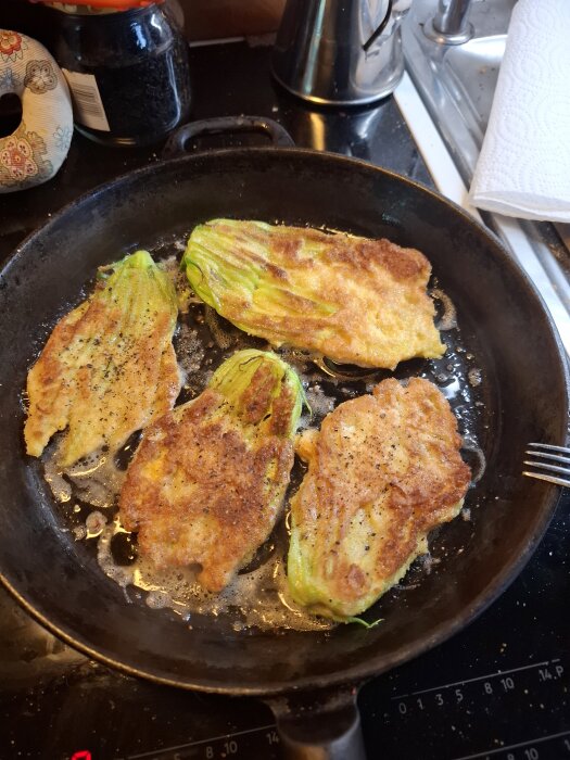 Fyra panerade och stekta zucchiniblommor i en stekpanna, placerad på en spis med en tekanna och hushållspapper i bakgrunden.