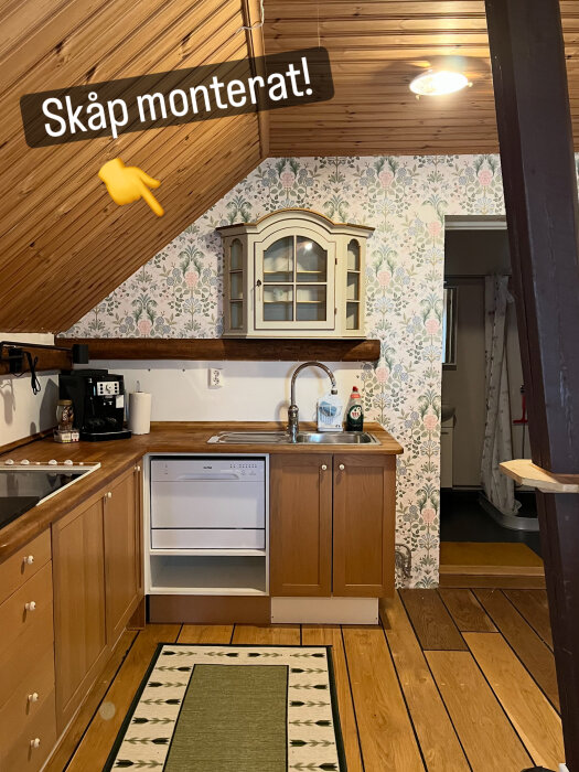 Kök med nytt monterat vitrinskåp ovanför diskbänken, träpanel i taket och mönstrad tapet på väggarna, diskmaskin saknas i skåpsutrymme.