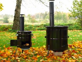Två utomhus värmekaminer på en gräsmatta omgiven av höstlöv, en med pipor för rökutsläpp, används för vattenuppvärmning i trädgården.