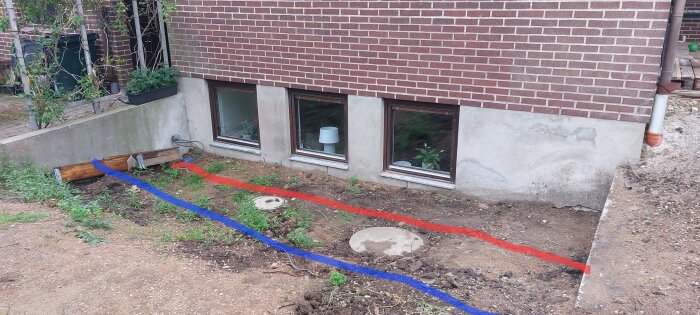 Slänt mot husgrund med markeringar i blått och rött där nya murar kan byggas. Två källarfönster och två brunnar synliga.