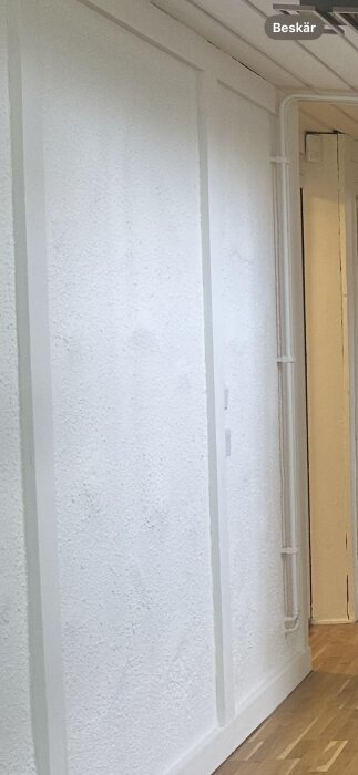 Strukturerad vit vägg med listverk i ett rum, träpanel i taket, trägolv, byggår 1974, möjligt behov av slätning och kontroll för asbest.