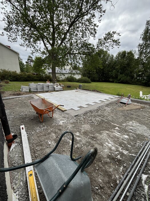 Byggprojekt i trädgård, 20 av 90 kvm klart, med verktyg som skottkärra, rätskiva och plattor.
