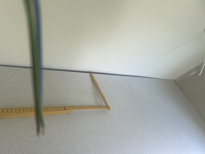 Mätsticka ligger på bänkskiva mot en vit vägg med en smal glipa bakom skivan. En kabel syns ovanför, hänvisar till köksrenovering.