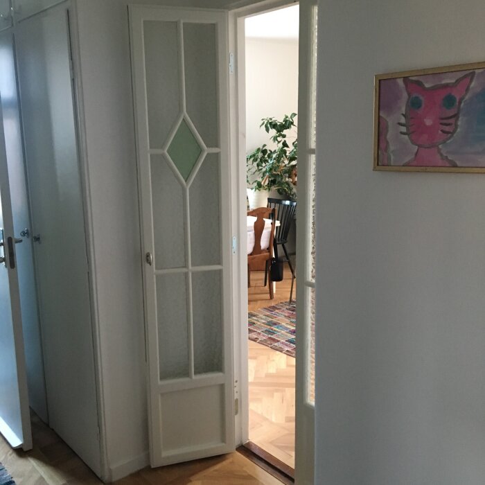 Minimal pardörr med glasdetaljer i en hall, dörren öppnas mot vardagsrummet, en tavla med rosa katthuvud på väggen.