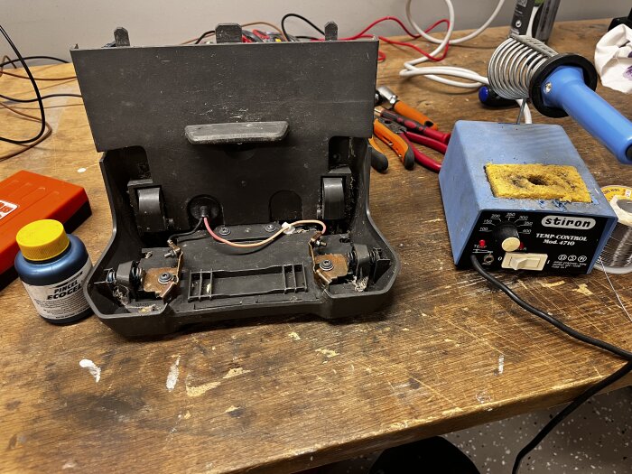 Inre del av en batteriladdare med trasiga kablar, lödstation till höger, verktyg och rengöringsmedel på en arbetsbänk.