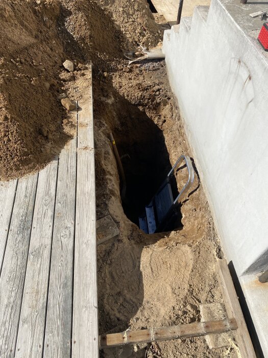 Grävt hål intill trätrall med synligt dräneringsrör och stege, omgiven av sand och jord.