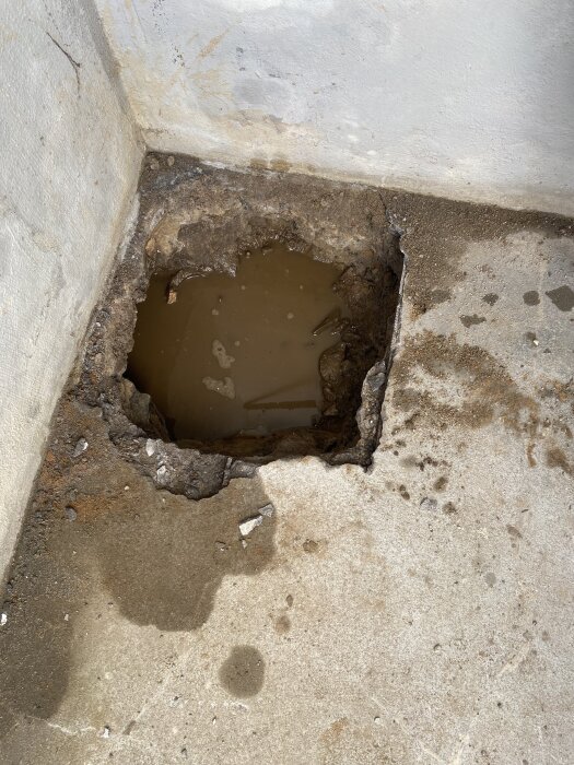 Hål i betonggolv med vatten och blottlagd jord vid dräneringsprojekt under trädäck.