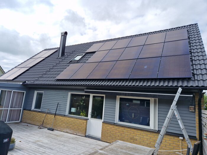 Solpaneler installerade på ett tak med betongpannor och takkrok. En stege lutar mot huset och terrassen syns i förgrunden.