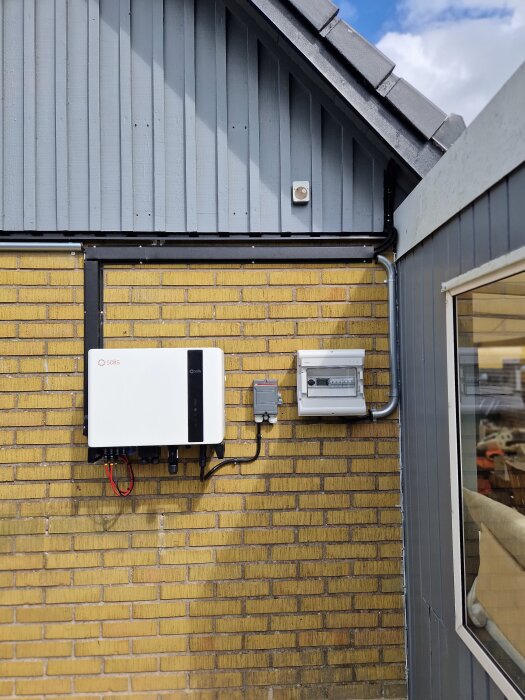 Solcellsinstallation på väggen av ett hus med gul tegel och grå panel. En växelriktare och elcentral är monterade bredvid varandra.