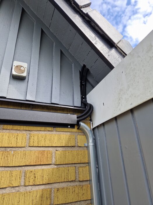 Exteriör av byggnad med gul tegelvägg och grå träpanel, visar rör och elektrisk kabeldragning längs väggen samt anslutning vid hörnet av taket.
