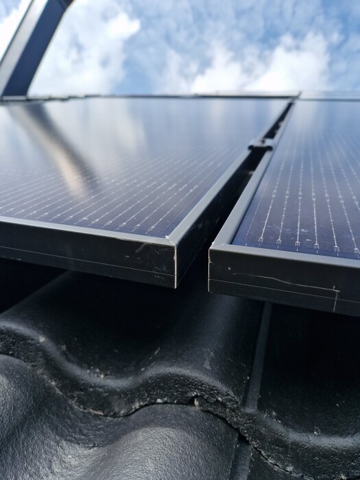 Närbild av solpaneler monterade på tak med takpannor och synliga springor mellan paneler och pannor, molnig himmel i bakgrunden.