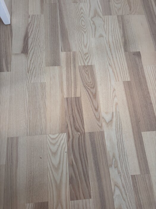 Parkettgolv av vitlaserad ask med 3-stavsmönster, mattlackad yta och olika längder på stavarna. Golvet har varierande träådringar och färgtoner.