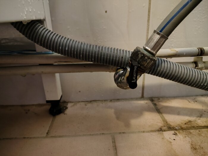 Närbild på en kulventil ansluten till en slang under en tvättmaskin i en tvättstuga, med fukt och smuts på golvet.
