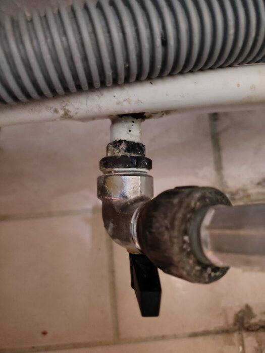 Närbild på kulventil och vattenkoppling med plastslang i tvättstuga, belägen på klinkergolv med vita fogar och synlig spricka i slangen.