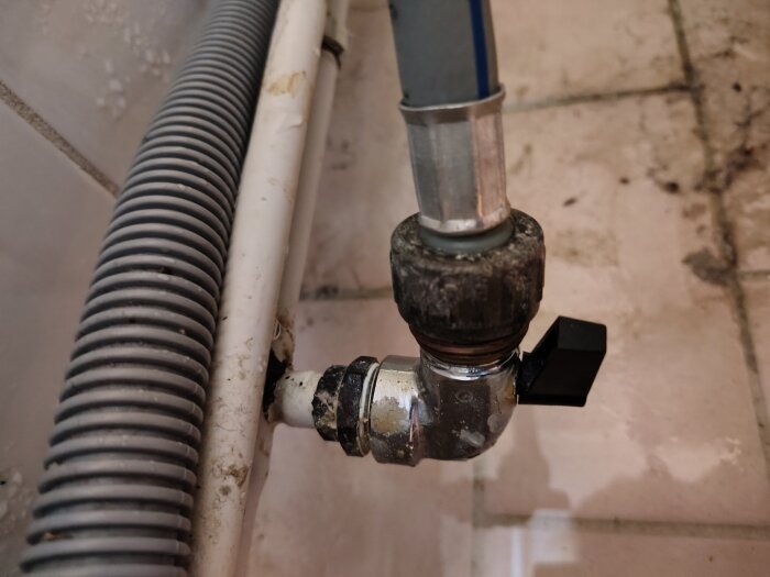 Kulventil och koppling med slang till tvättmaskin i tvättstuga, synlig spricka på slangen och märken av vattenläckage på kakelgolvet.