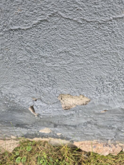 Färgen på en grå vägg har börjat flagna och det syns skador i putsen, med gräs vid basen av väggen.