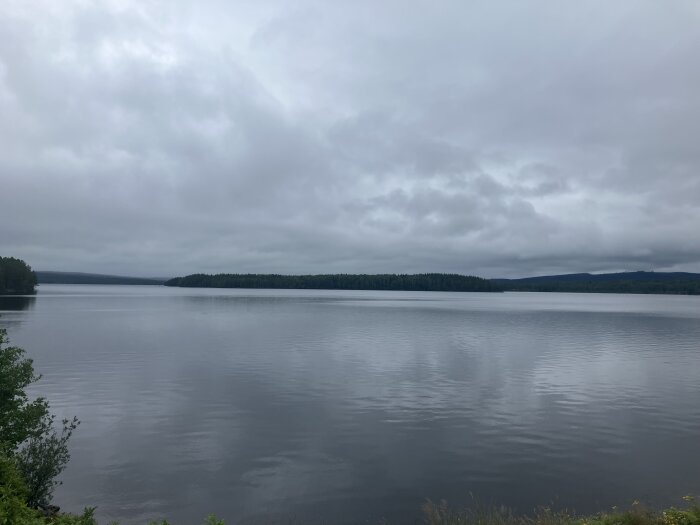 Molnig himmel över en spegelblank sjö med skogsklädda stränder i bakgrunden.