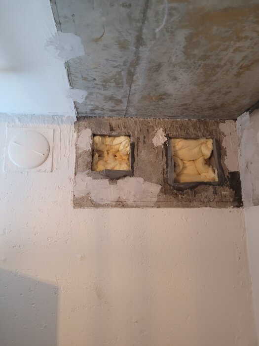 Två kvadratiska ventilationshål i väggen fyllda med gul isolering, väntar på att tätas med fogskum för efterföljande putsning och målning.