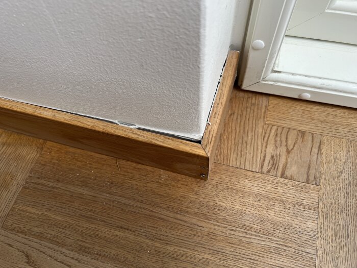 Trälist delvis lossnad från vit vägg i hörn med parkettgolv.
