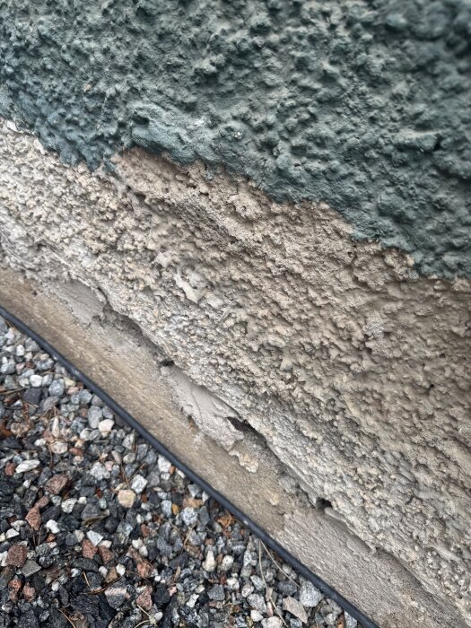 Närbild av sliten fasad med skrovlig yta, delvis målad i grönt, och makadam vid basen.