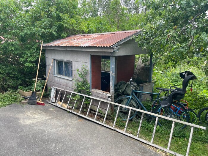 Fallfärdig lekstuga med eternitväggar, stege lutad mot väggen, cyklar och trädgårdsredskap. Omgiven av grönska, behöver rivas.