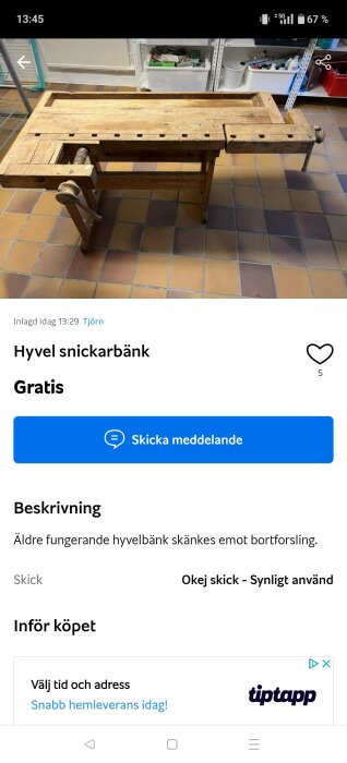 Äldre träbearbetningsbänk med vev och bänkyta, placerad på ett kaklat golv i ett verkstadsutrymme.