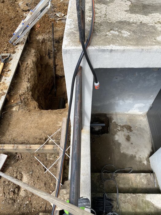 Grävprojekt med synliga rör, jord och betongyta, det gamla röret syns i bakgrunden. Stege och verktyg är också med i bilden.