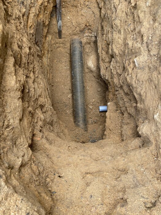 Grävd grop med exponerat gammalt svart dräneringsrör och vitt 50 mm rör i jord.