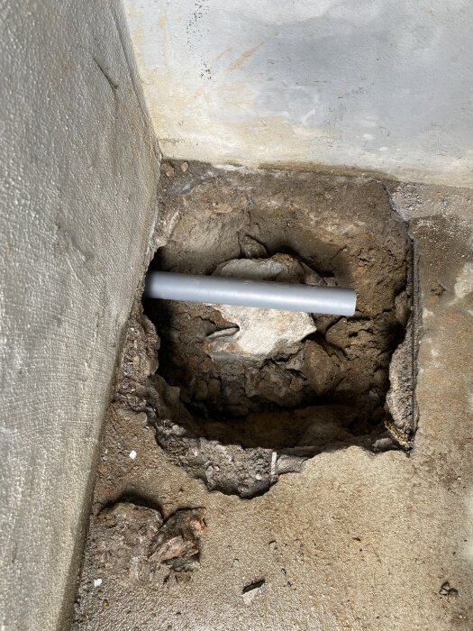 Ett grått rör syns liggande i en rektangulär hålighet i en betonggolvyta, med mörk jord och sten runtomkring.