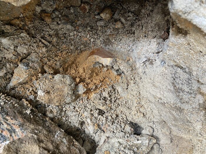 Bild av en grop med jord och sten där ett brunt rör är synligt, omgivet av grå och brun jord samt stenar.