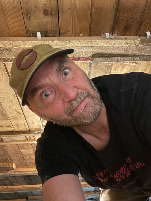 Person inspekterar trätak i låg källare under ett fritidshus. Bär keps och har ögonkontakt med kameran.
