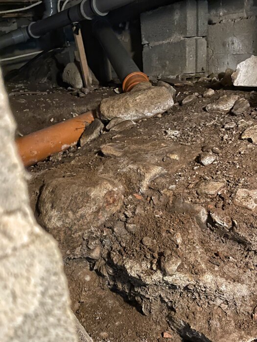 Utrymme under ett fritidshus med synliga rör, jord och stenar, samt del av murad vägg. Dränering och rörsystem kontrolleras.