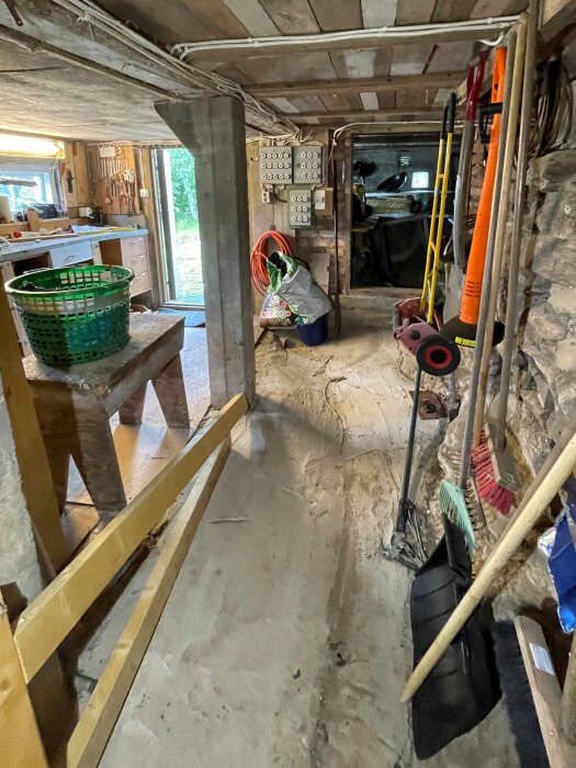 Källarutrymme under ett fritidshus med verktyg, rör, och synliga kablar. Låg takhöjd, stenar och brädor ses längs väggen, med trädgårdsredskap i förgrunden.