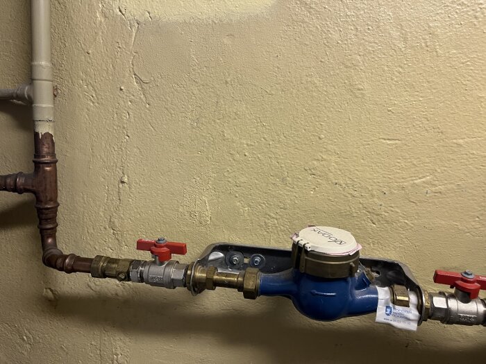Vattenmätare med kopparrör som minskar i diameter från 28 mm till 22 mm efter mätaren, synliga ventiler och anslutningar på ljus vägg.
